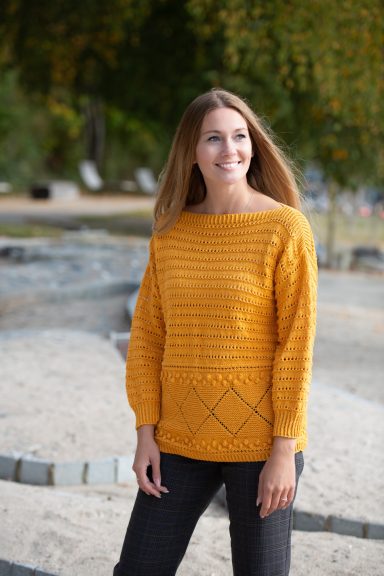 Femme souriante portant un pull en laine couleur moutarde fait main, devant un décor extérieur.
