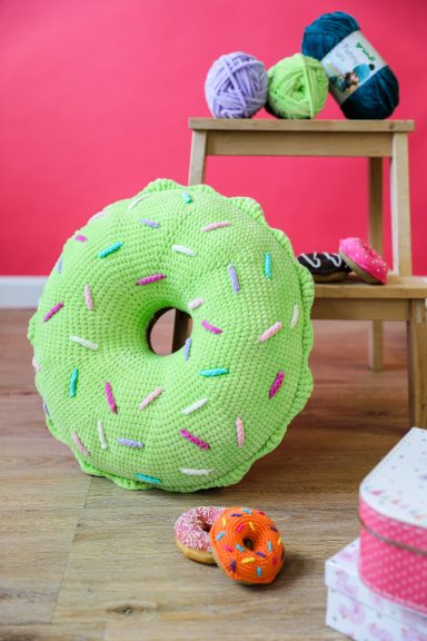 Coussin en forme de beignet vert avec des sprinkles colorés, décoré sur fond rose.